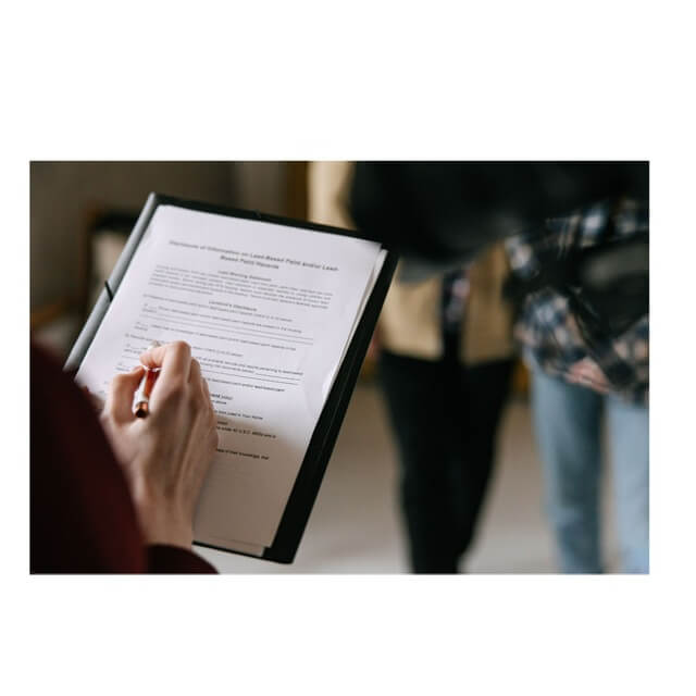 Close-up of a hand writing on a form held on a clipboard as people stand in the background in a meeting setting.