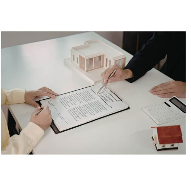 Hands signing a Real Estate Contract on a white table, with a wooden house model and a calculator nearby.
