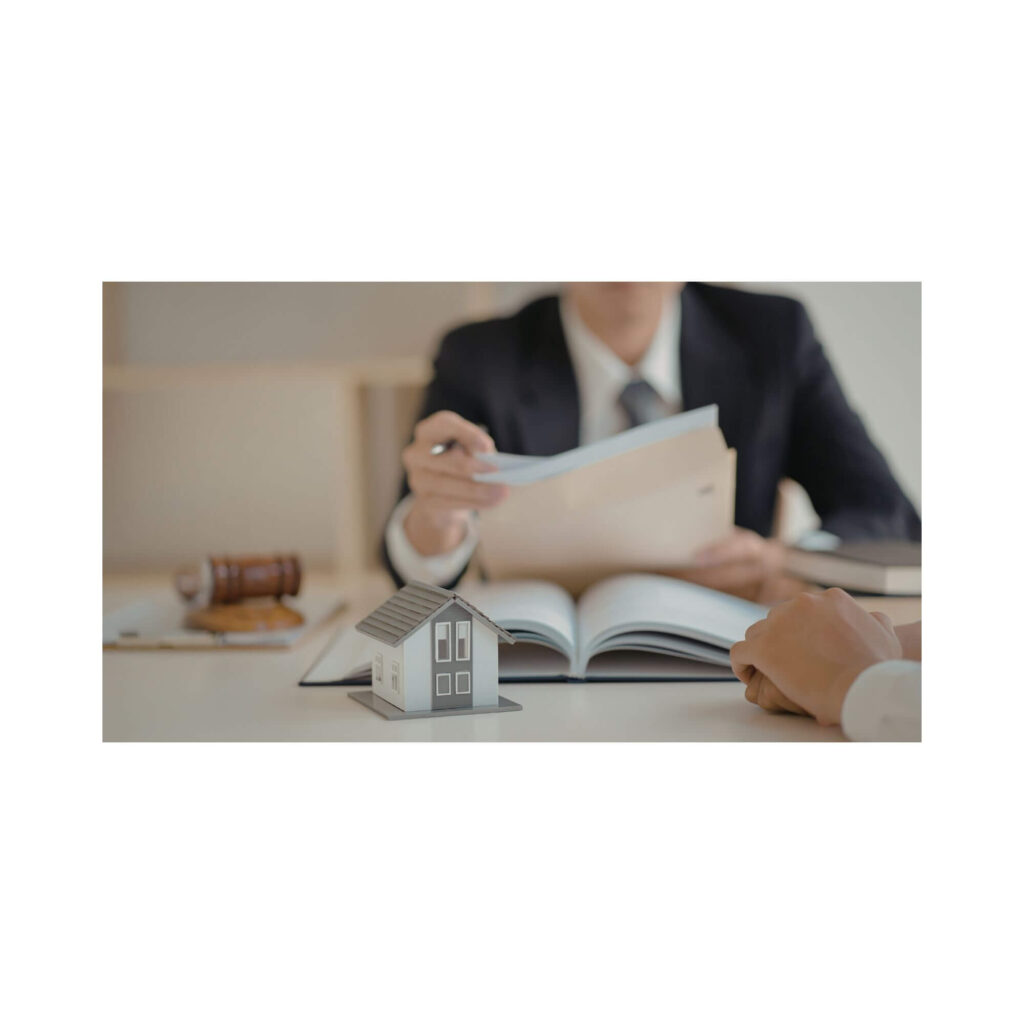A person in a suit reviewing property documents across a table from another person’s hand, with a model house between them